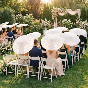 May include: Outdoor wedding ceremony with guests seated in white folding chairs. Attendees hold white paper parasols. The setting is a lush green lawn with floral arrangements and a decorated archway.