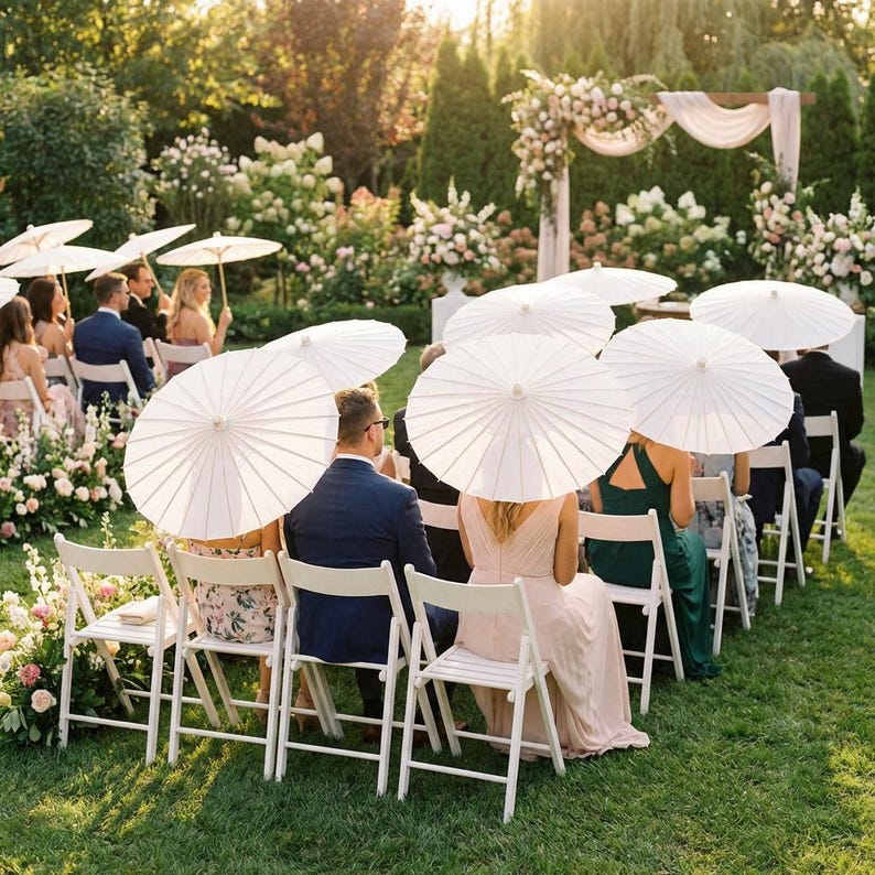 May include: Outdoor wedding ceremony with guests seated in white folding chairs. Attendees hold white paper parasols. The setting is a lush green lawn with floral arrangements and a decorated archway.