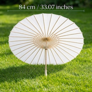 May include: A white paper parasol with a bamboo frame, measuring 33.07 inches in diameter, is open on a bed of green grass. The parasol's ribs radiate from a central point, creating a sunburst pattern. The handle is made of wood.