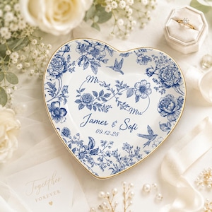 Op de afbeelding: Hartvormige keramische schaal met een blauw bloemmotief en gouden rand. De schaal is gepersonaliseerd met de namen "James & Sofi" en de datum "09.12.25". Decoratieve elementen zijn bloemen en vogels.