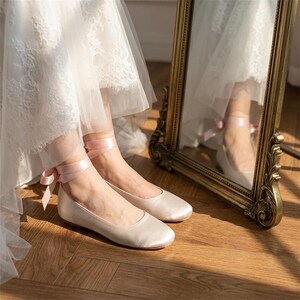 Bridal ballet flats with ribbon ties in soft blush satin, comfortable wedding shoes for bride, minimalist flat shoes for ceremony and reception, elegant lightweight bridal footwear on wooden floor