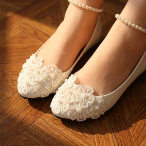 Ivory bridal lace flats with pearl details on feet, elegant wedding ballet shoes with floral lace design, comfortable bride shoes for ceremony and reception on wooden floor