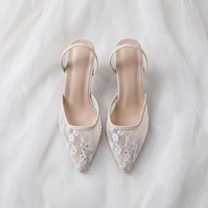 Elegant lace slingback heels with pointed toe and sculpted heel, available in white and black colors, styled in romantic bridal setting with pearls, lace fabric, and floral details for wedding or special occasion wear.