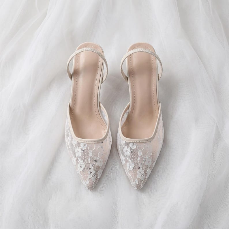 Elegant lace slingback heels with pointed toe and sculpted heel, available in white and black colors, styled in romantic bridal setting with pearls, lace fabric, and floral details for wedding or special occasion wear.