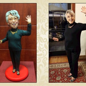 May include: A woman in a black turtleneck sweater and black pants is standing in front of a window. She is waving with her right hand. A figurine of the woman in the same pose is standing on a red circular base.