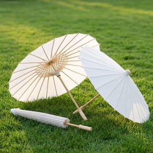 Three white paper parasols with wooden handles rest on green grass—two are open and lying at angles, showing the delicate rib structure, while one is closed and placed in front of them.