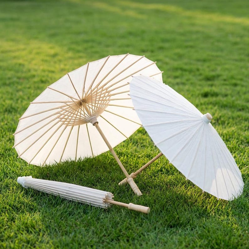 Three white paper parasols with wooden handles rest on green grass—two are open and lying at angles, showing the delicate rib structure, while one is closed and placed in front of them.