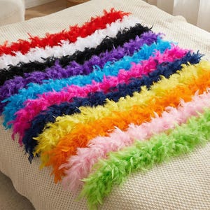 Op de afbeelding: Een verzameling kleurrijke verenboa's, gerangschikt in parallelle rijen. De boa's tonen een levendig spectrum aan kleuren, waaronder rood, wit, zwart, paars, blauw, roze, marineblauw, geel, oranje, lichtroze en groen. Gepresenteerd op een beige, getextureerd oppervlak.