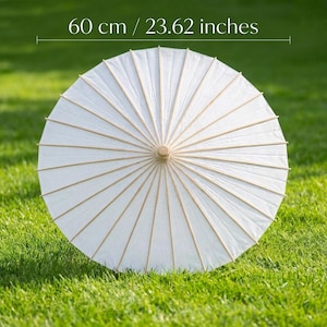 May include: A white paper parasol, 23.62 inches in diameter, with a wooden handle and ribs. The parasol is open and resting on green grass. The parasol is a traditional design, often used for sun protection or decoration.