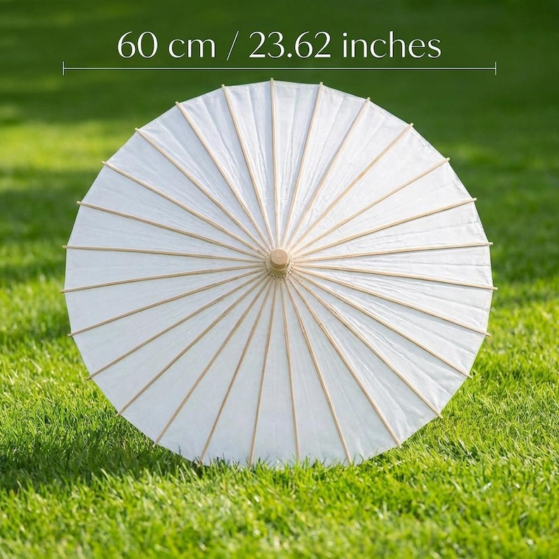 May include: A white paper parasol, 23.62 inches in diameter, with a wooden handle and ribs. The parasol is open and resting on green grass. The parasol is a traditional design, often used for sun protection or decoration.