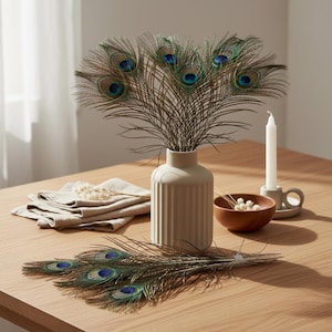 Può includere: Un vaso beige con linee verticali contiene piume di pavone con segni oculari blu e verdi. Un mazzo di piume di pavone è appoggiato su un tavolo di legno, insieme a una candela in un portacandele grigio, una piccola ciotola di legno e un tovagliolo di lino beige piegato.