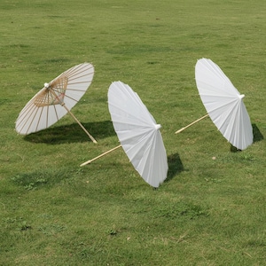 May include: Three white paper parasols with bamboo handles are arranged on a green grassy surface. The parasols are open, displaying their pleated white paper canopies. One parasol has a visible bamboo frame.