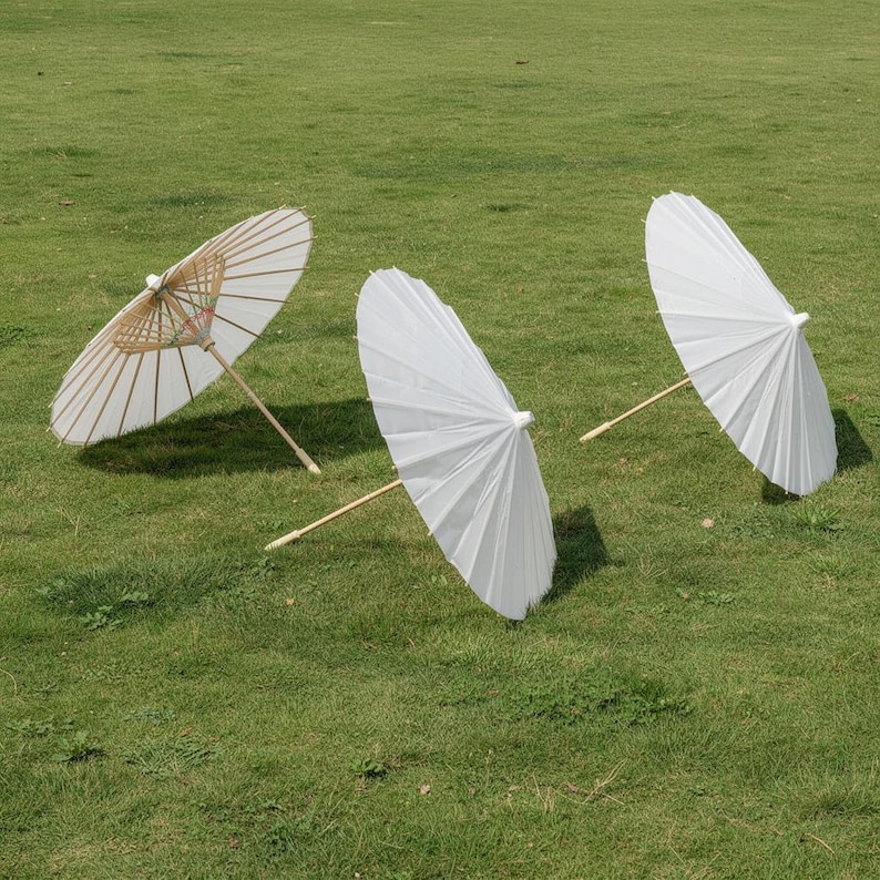 May include: Three white paper parasols with bamboo handles are arranged on a green grassy surface. The parasols are open, displaying their pleated white paper canopies. One parasol has a visible bamboo frame.