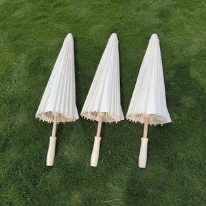 May include: Three closed white paper parasols with light wood handles are arranged on green grass. The parasols have a pleated design and a scalloped edge. The handles are a light wood color.