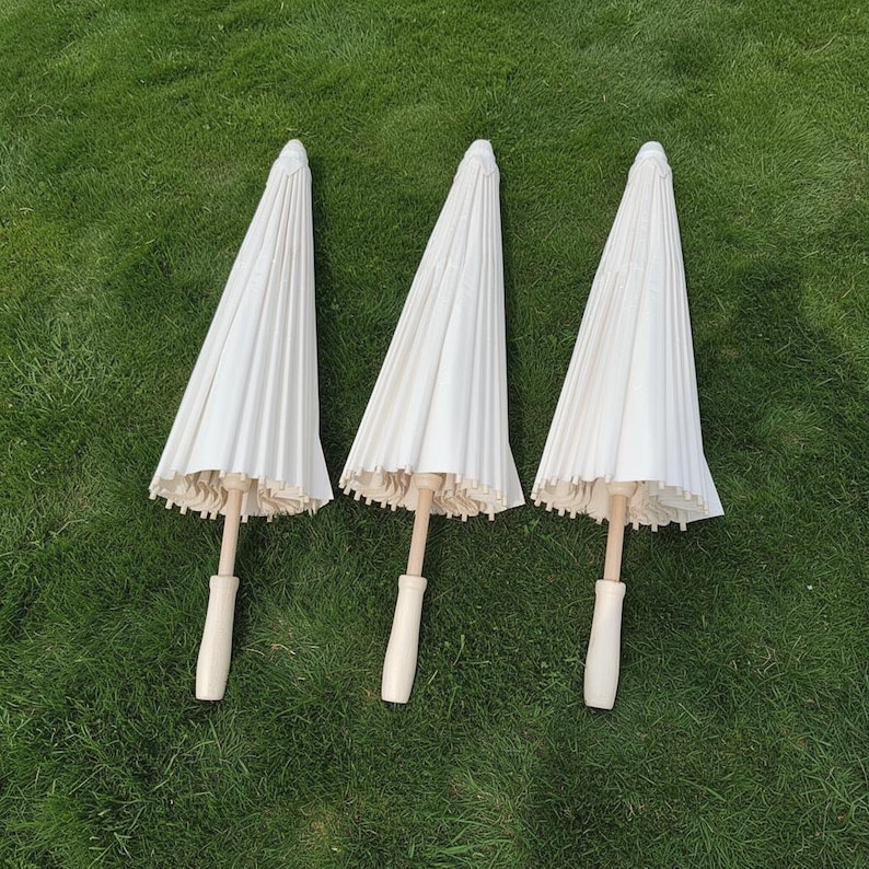 May include: Three closed white paper parasols with light wood handles are arranged on green grass. The parasols have a pleated design and a scalloped edge. The handles are a light wood color.