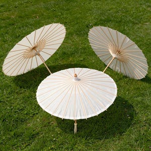May include: Three open, off-white paper parasols with wooden handles and ribs are arranged on a bed of green grass. The parasols are circular, with a tassel hanging from the bottom of one.