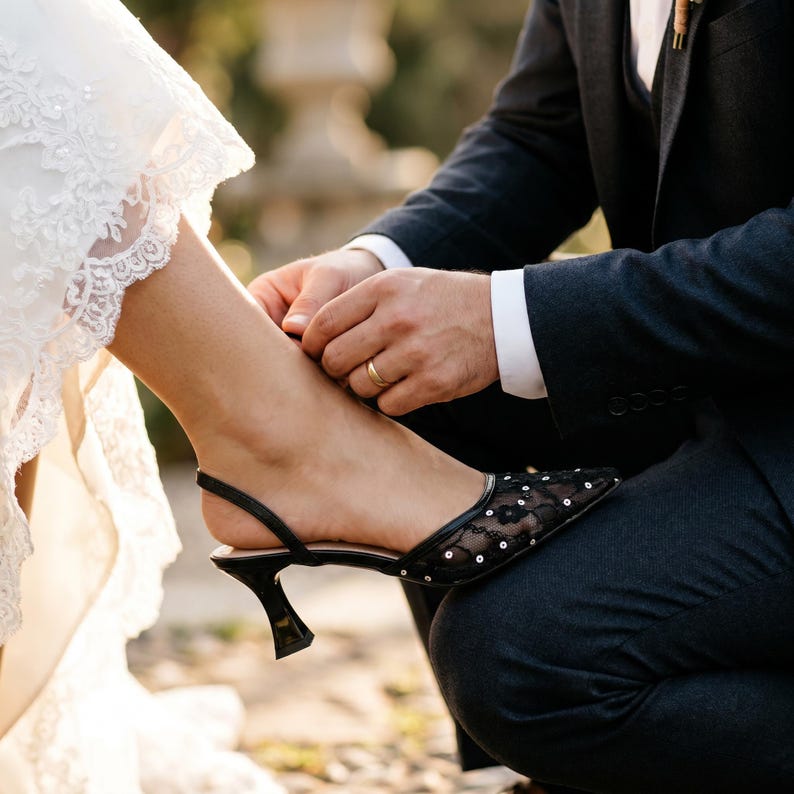 Elegant lace slingback heels with pointed toe and sculpted heel, available in white and black colors, styled in romantic bridal setting with pearls, lace fabric, and floral details for wedding or special occasion wear.