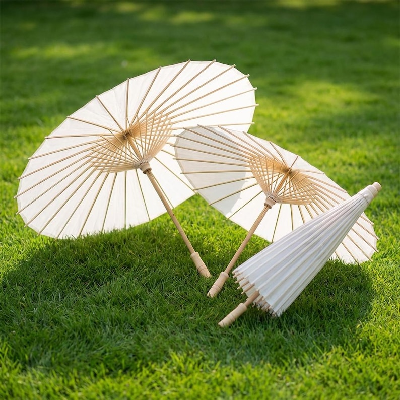 May include: Three white paper parasols with bamboo handles are arranged on a bed of green grass. Two are open, displaying their fan-like structure, while the third is closed. The parasols are a light cream color.