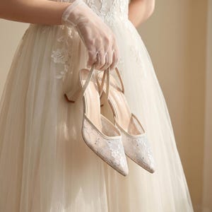 Puede incluir: Un par de zapatos de tacón de encaje marfil para bodas con punta y tacón pequeño. Los zapatos los sostiene una persona con un vestido blanco y guantes transparentes. Diseño delicado y femenino.