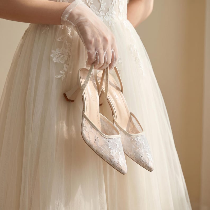 Elegant lace slingback heels with pointed toe and sculpted heel, available in white and black colors, styled in romantic bridal setting with pearls, lace fabric, and floral details for wedding or special occasion wear.
