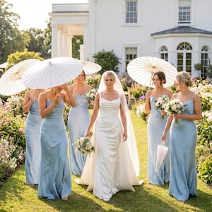 May include: A bride in a white satin wedding gown with a veil walks down a grassy path, followed by bridesmaids in light blue dresses. The bridesmaids carry bouquets and parasols. A large white house is in the background.