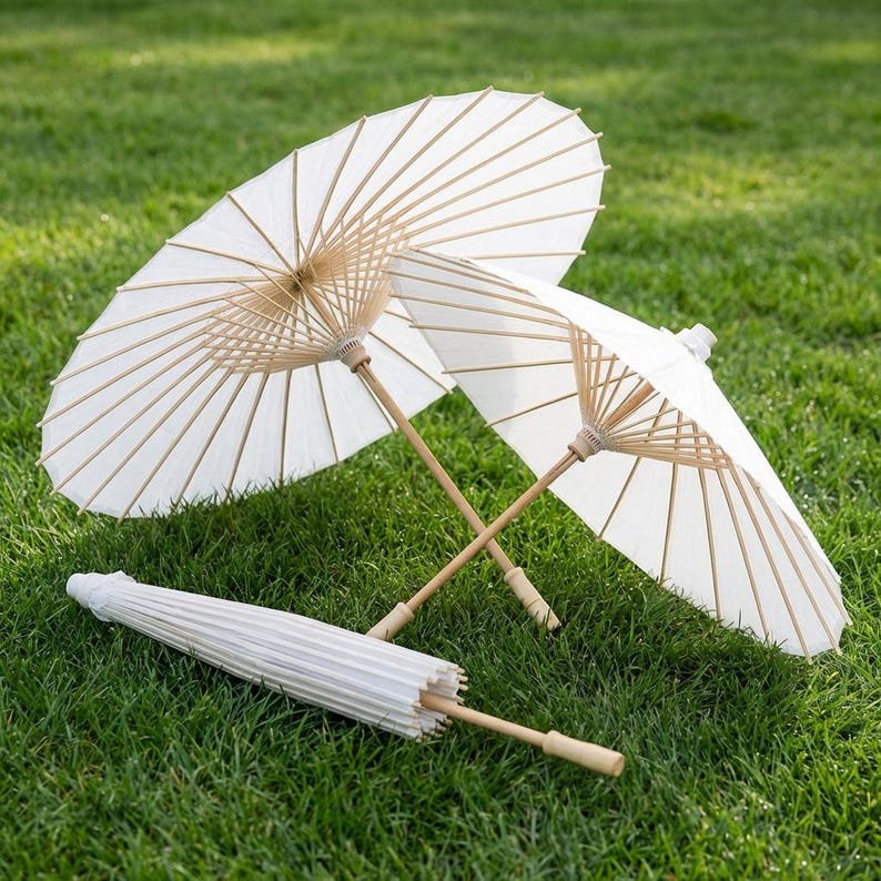 May include: Three white paper parasols with bamboo handles are arranged on green grass. Two are open, displaying their delicate, fan-like structure, while the third is closed. The parasols are ideal for outdoor events or decorative purposes.