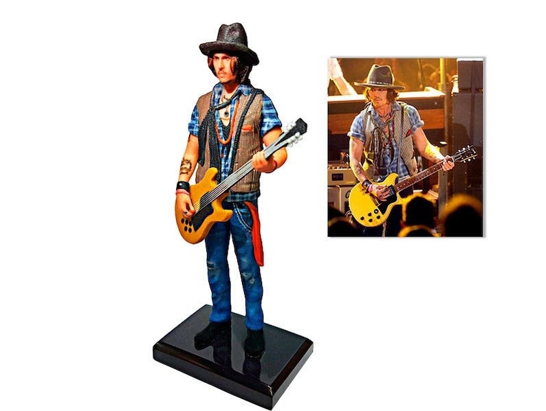 May include: A figurine of a musician playing a yellow electric guitar. The figurine is wearing a brown hat, a blue and white plaid shirt, a brown vest, and blue jeans. The figurine is standing on a black base.