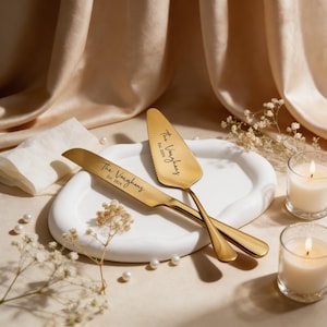 May include: Gold-colored cake knife and server set with the inscription "The Vaughans Est. 2024" on each piece. The set rests on a white, oval-shaped plate, with lit candles and pearls in the background. The overall theme is wedding or celebration.