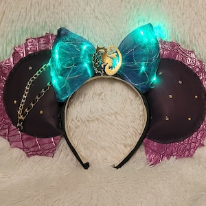 May include: Black and purple mouse ears headband with a teal bow and gold dragon emblem. The ears have a chain and studded accents, with a textured purple trim. The bow lights up with teal lights.