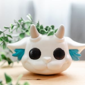 May include: A white ceramic planter shaped like a cartoon dragon with black eyes, small horns, and blue wings. The planter is filled with green plants, creating a whimsical and decorative piece for home decor.