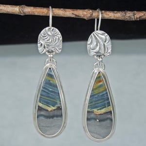 May include: A pair of silver drop earrings featuring a teardrop-shaped stone with horizontal bands of blue, yellow, and gray. Each earring has a textured, semi-circular silver top piece with an embossed leaf-like pattern and a silver hook wire.