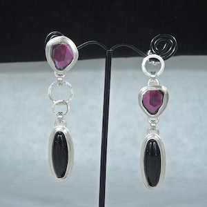 May include: A pair of dangling earrings with a silver setting. Each earring features a dark pink, irregularly shaped stone at the top, connected by silver rings to a black, oval-shaped stone at the bottom. The earrings are displayed on a black stand.