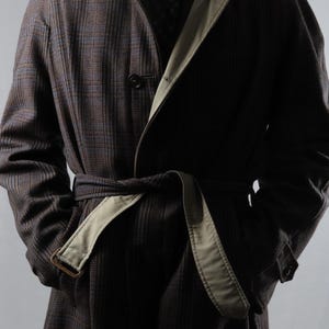 Peut inclure: Un long manteau marron foncé avec un motif à carreaux subtil, une doublure de revers beige et une ceinture à nouer assortie à la taille. Le manteau est porté sur une chemise blanche à col et une cravate sombre à motifs, avec des boutons visibles.