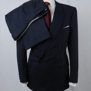 Puede incluir: Un traje cruzado de color azul marino con rayas diplomáticas y pantalones a juego colocados sobre el hombro. El conjunto incluye una camisa blanca de cuello rígido, una corbata roja oscura lisa y un pañuelo blanco en el bolsillo del pecho.