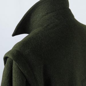 Könnte beinhalten: Nahaufnahme eines dunkelgrünen Wollmantels. Der Mantel hat einen breiten, spitz zulaufenden Kragen und ein strukturiertes Gewebe. Der Schulterbereich ist sichtbar und zeigt die Konstruktion des Mantels. Der Gesamteindruck ist klassisch und maßgeschneidert.
