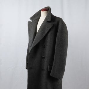 Peut inclure: Un manteau en laine gris anthracite à double boutonnage avec un large revers cranté, des manches longues et trois boutons foncés visibles sur le devant. Le manteau est présenté sur un mannequin, révélant une silhouette structurée et un col classique.