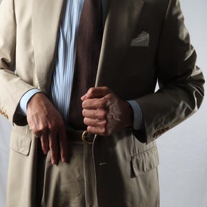Peut inclure: Un costume beige composé d'une veste et d'un pantalon assorti, porté avec une chemise à rayures bleu clair et blanches, une cravate texturée marron foncé et une pochette blanche. La veste présente des revers crantés et des boutons aux poignets.