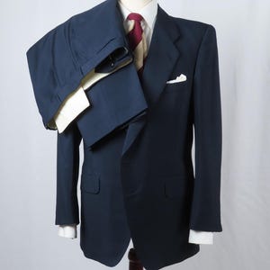 Op de afbeelding: Een marineblauw pak op een mannequin. Het pak bestaat uit een jasje met een wit pochet en bijpassende broek. Een wit overhemd en een stropdas met patroon maken de outfit compleet. Het pak wordt gepresenteerd op een houten standaard.