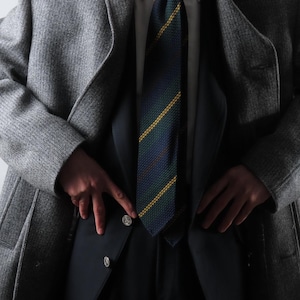 May include: A gray wool overcoat worn over a navy suit, white shirt, and patterned tie. The tie features a diagonal pattern of navy, green, and yellow stripes. The suit jacket has two silver buttons.