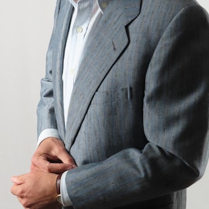 May include: A light blue blazer with a textured weave, worn over a striped shirt. The jacket has a notched lapel and a single-breasted design. The sleeves are cuffed, showing a brown leather watch strap.