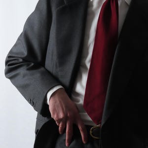 Könnte beinhalten: Ein dunkelgrauer Anzug mit passender Jacke und Hose. Eine weinrote Krawatte wird mit einem weißen Hemd kombiniert. Der Anzug hat eine klassische, maßgeschneiderte Passform und einen schwarzen Gürtel mit goldener Schnalle.