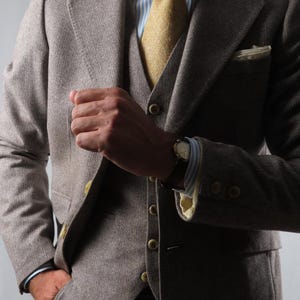 Puede incluir: Un traje de tres piezas en tela marrón texturizada que incluye una chaqueta, un chaleco abotonado y una camisa de rayas azul claro y blancas. Se lleva una corbata de color amarillo mostaza liso y un pañuelo de bolsillo blanco en la chaqueta.
