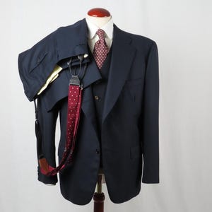 Puede incluir: Un traje azul marino exhibido en un maniquí. El conjunto incluye una chaqueta, un chaleco y un pantalón, combinados con una camisa blanca con cuello y una corbata roja estampada. También se ven tirantes y una corbata roja de lunares.