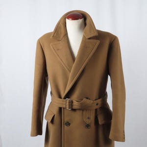 Puede incluir: Un abrigo marrón de doble botonadura con cuello con solapas, cinturón y botones. El abrigo se exhibe en un maniquí. Tiene un cinturón con hebilla plateada y dos bolsillos delanteros.