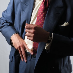 Könnte beinhalten: Ein marineblaues Sakko über einem cremefarbenen Hemd. Das Outfit umfasst eine dunkelrote Krawatte mit einem sich wiederholenden gold-blauen Fleur-de-Lis-Muster, ein weißes Einstecktuch sowie einen schwarzen Ledergürtel mit einer goldfarbenen Uhr.