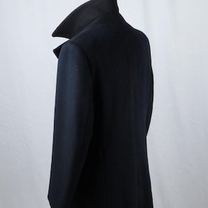 May include: A dark navy blue coat featuring a high, structured collar and long sleeves. The garment is shown from a side-back angle, displaying a clean, tailored silhouette with a smooth texture and a single vertical seam along the back panel.