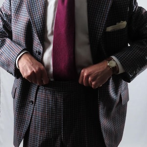 May include: A patterned suit with a burgundy tie. The suit jacket has a pocket square and a watch is visible on the wrist. The suit is a mix of blue, brown, and gray. The shirt is white.