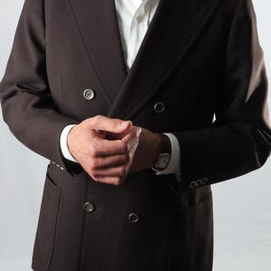 May include: A dark brown double-breasted blazer with a notched lapel and two rows of buttons. The blazer has two front pockets and is worn over a white dress shirt. A gold watch is visible on the wrist.