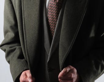 Op de afbeelding: Een donkergroene overjas gedragen over een grijs vest en een wit overhemd met kraag. Een stropdas met patronen met rode en witte accenten is zichtbaar. De jas is dichtgeknoopt en de persoon houdt de revers vast.