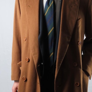 May include: A double-breasted, camel-colored overcoat with large, round buttons. The coat is worn over a dark vest, a patterned tie with green and yellow stripes, and a white collared shirt. The coat has a classic, tailored design.